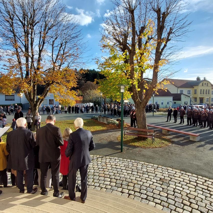 Eine Menschenmenge versammelt sich an einem sonnigen Tag in einem Park. Sie sind in formeller Kleidung gekleidet, und einige tragen Hüte. Es gibt Bäume mit gelben Blättern, Bänke und Straßenlaternen. In der Ferne stehen Gebäude.