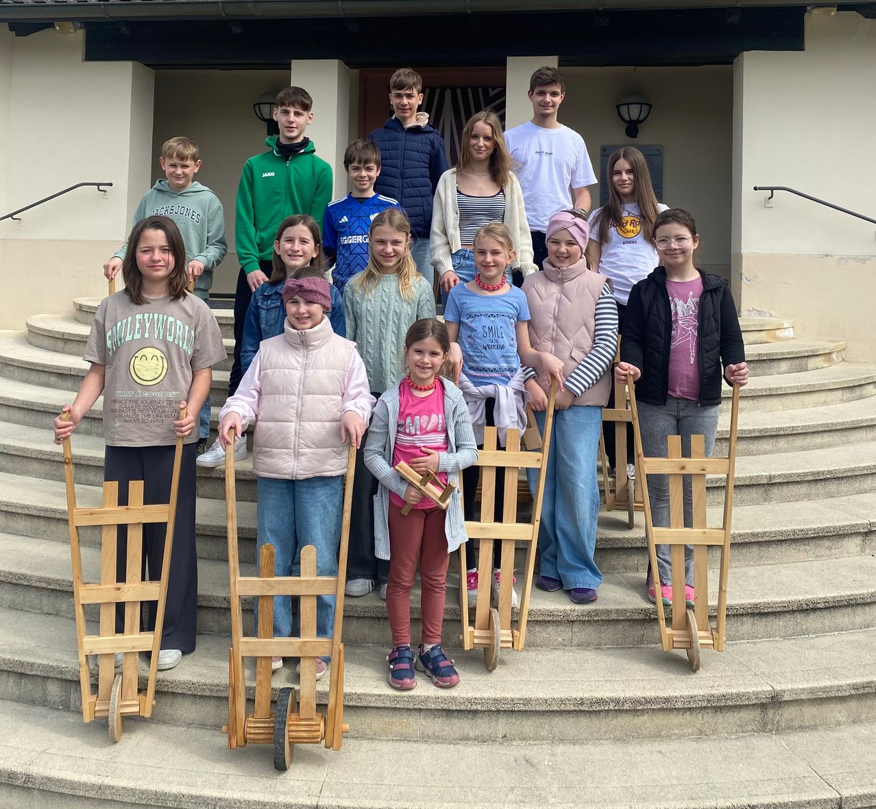 A group of young people are posing with wooden sleds on a set of stairs. Some are wearing jackets, while others have beanies on. They are smiling for the camera.