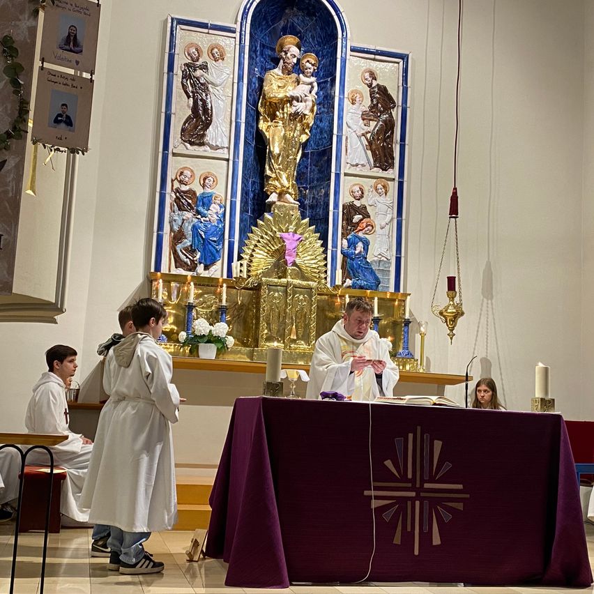 A priest is leading a service in a church with three altar boys, candles, and a golden statue in the background.