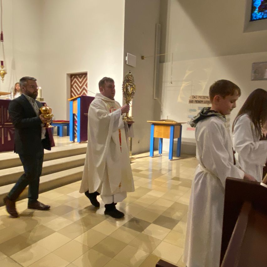 A priest and acolytes hold sacred objects inside a church. A man in a suit holds a golden chalice.