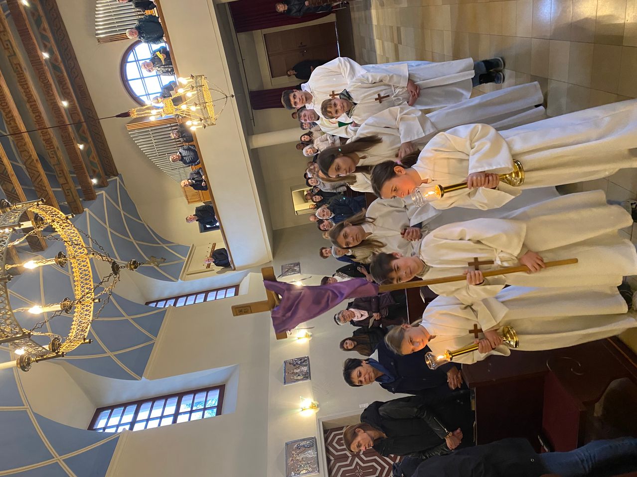 Group of children in white robes holding candles, possibly during a religious service in a church.