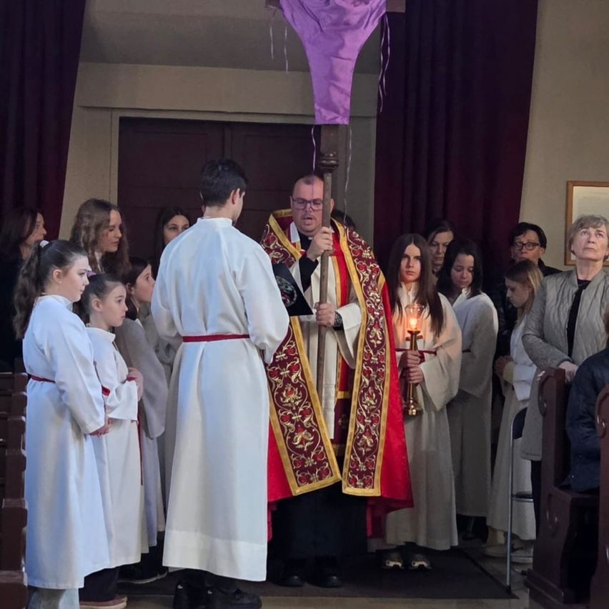 A priest in a red and gold robe stands before a group of young people holding candles in a church.