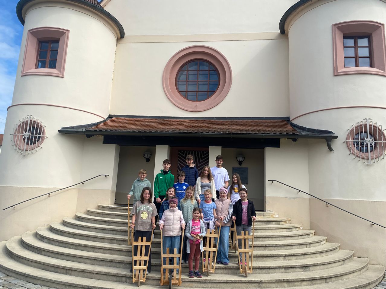 A group of children, wearing winter clothing, stand on the stairs in front of a building with a round window. They hold wooden sleds and pose for a photo.