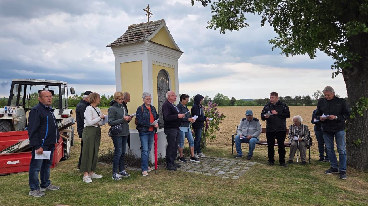Eine Gruppe von Menschen steht um eine Kapelle mit einem Kreuz oben herum, einige lesen von Papieren. Eine Person sitzt auf einer Bank, eine andere hält einen Regenschirm.