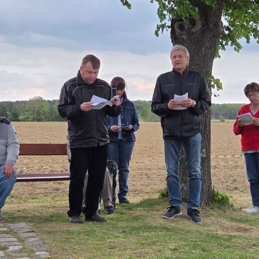 Fünf Menschen stehen in einem offenen Feld und lesen aus Papieren. Ein Baum rechts, eine Bank links. Bäume und ein bewölkter Himmel im Hintergrund.
