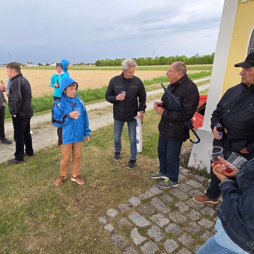 Eine Gruppe von Menschen steht auf einem Ziegelweg, einige halten Getränke. Ein Mann in einer blauen Jacke sticht hervor, während andere schwarze Jacken tragen. Ein Regenschirm ist geöffnet, und im Hintergrund zeigt sich ein grasiges Feld.