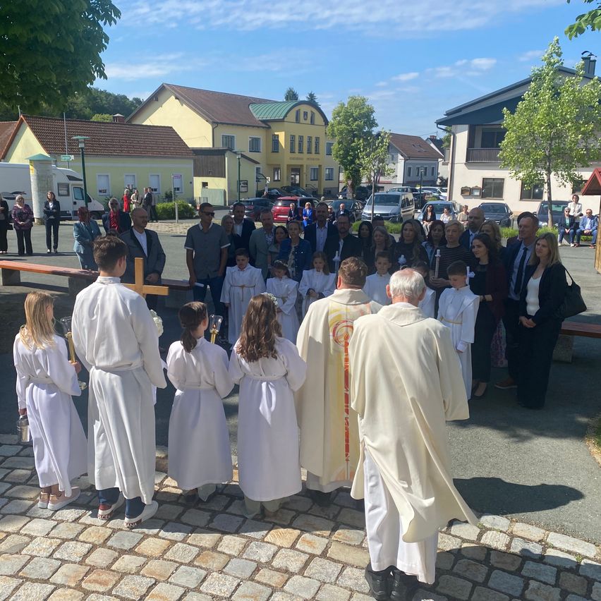 Eine Gruppe von Menschen versammelt sich für eine Zeremonie vor einer Kirche mit Gebäuden und Bäumen im Hintergrund.