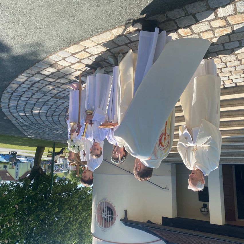Eine Gruppe von Priestern in weißen Roben steht auf Kopfsteinpflaster und hält Gegenstände. Der Boden ist reflektiert und das Gebäude dahinter hat ein Fenster mit Gitterstäben.