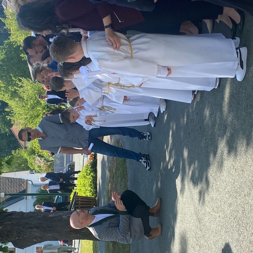 Eine Gruppe von Menschen, darunter Kinder in weißen Roben, steht im Kreis, wobei ein Mann in Anzug vor ihnen kniet. Sie scheinen an einer religiösen Zeremonie teilzunehmen.