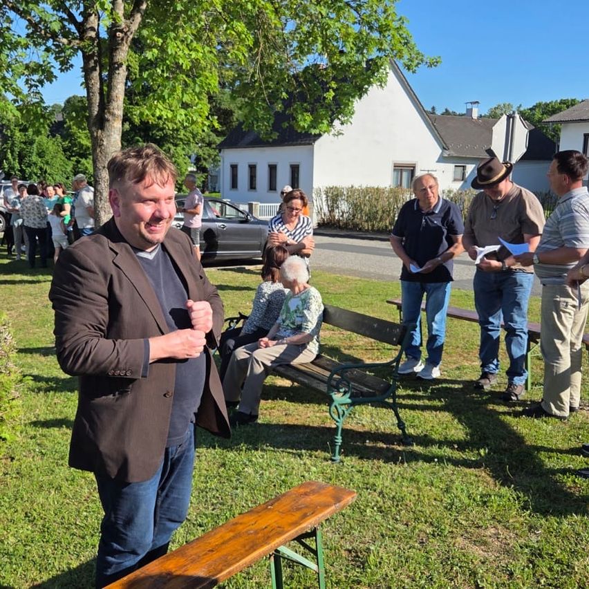 Eine Gruppe von Menschen versammelt sich in einem Park. Ein Mann steht vor einer Holzbank und lächelt. Hinter ihm sitzen andere auf einer Bank. Ein Auto ist auf der Straße geparkt.