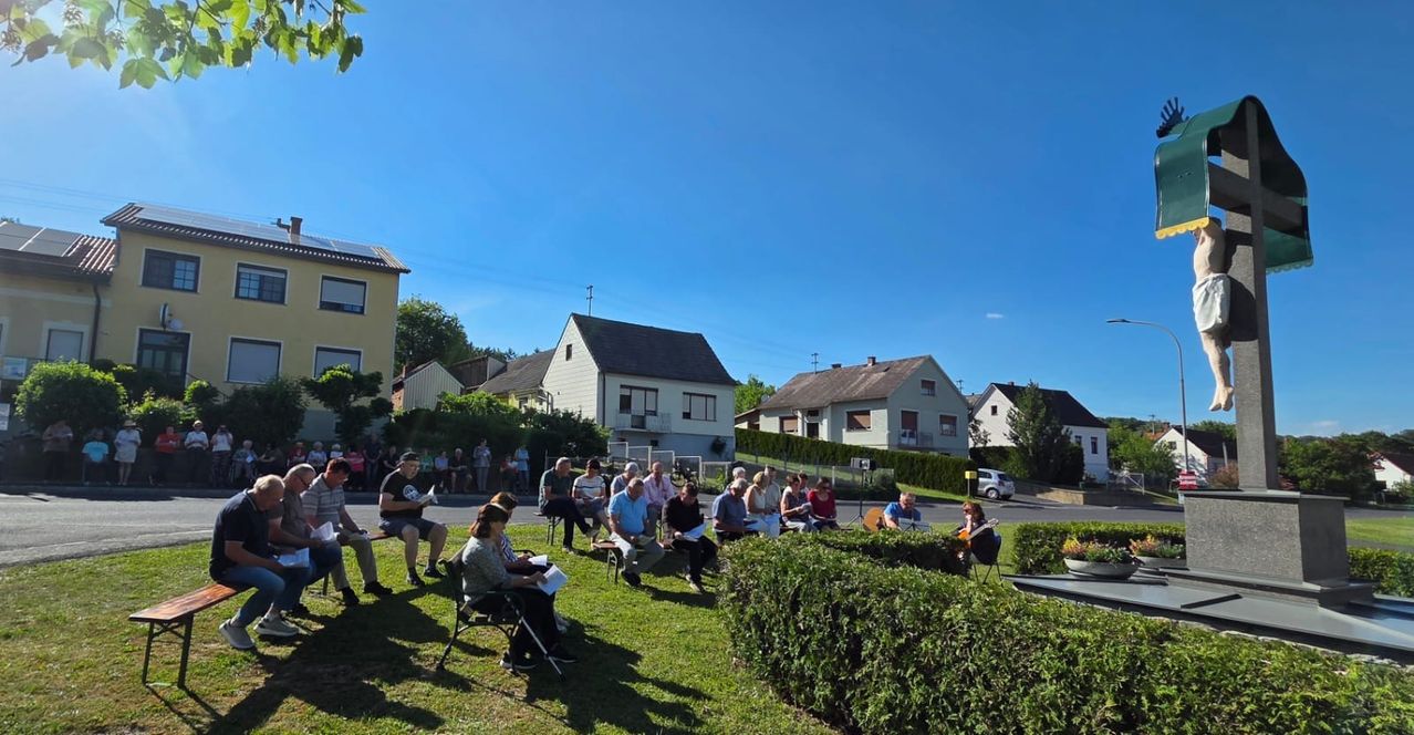 Eine Gruppe von Menschen versammelt sich auf einem Rasen, einige sitzen auf Stühlen, andere stehen, mit Häusern und Grün im Hintergrund. Eine Frau spielt Gitarre, während die Leute lesen und zuhören.