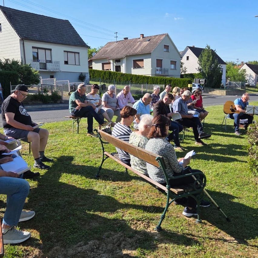 Eine Gruppe von Menschen sitzt auf Bänken in einem Grasbereich, einige lesen Bücher, während andere reden. Eine Person mit einer Gitarre steht. Dahinter sind Häuser und eine Straße.