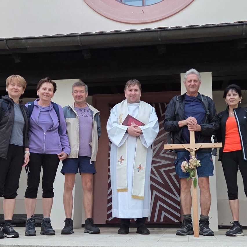 Eine Gruppe von Menschen, einschließlich eines Priesters, steht vor einer Kirche und lächelt für ein Foto. Der Priester hält ein Buch und ein Mann hält ein Kreuz.