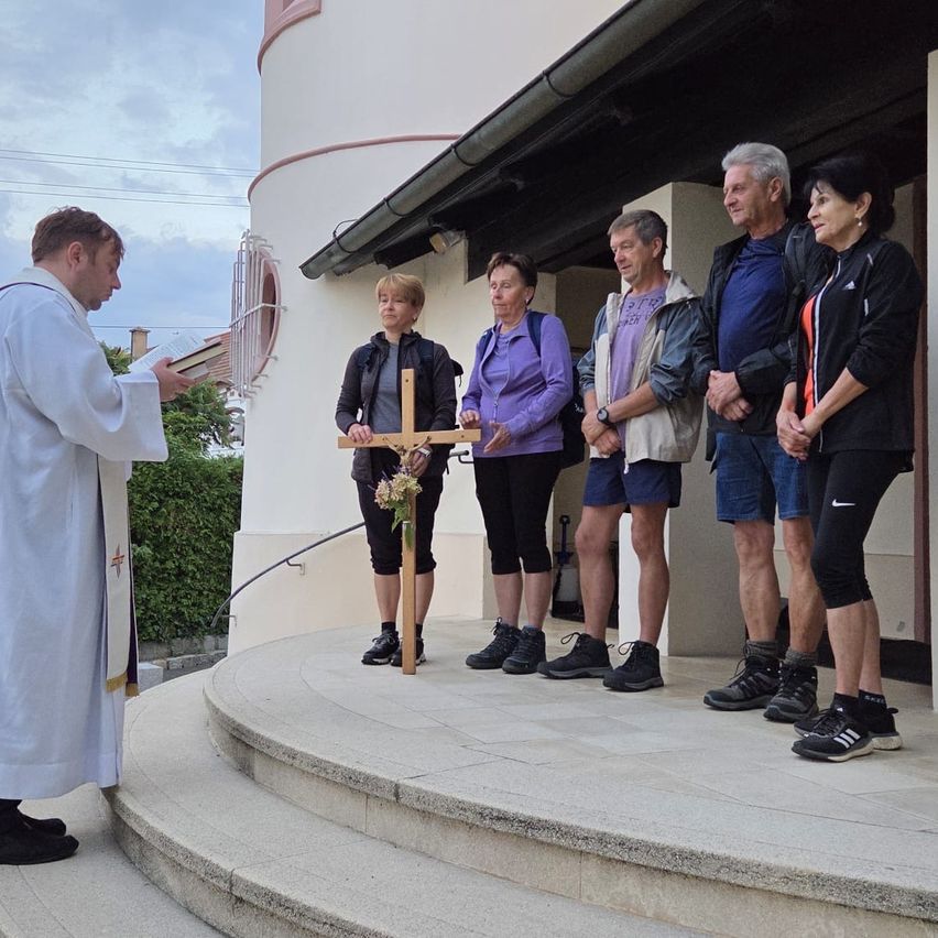 Ein Priester in einer weißen Robe leitet eine Gruppe von Menschen in einem Gebet vor einer Kirche an, wobei eine Frau ein Kreuz und Blumen hält.