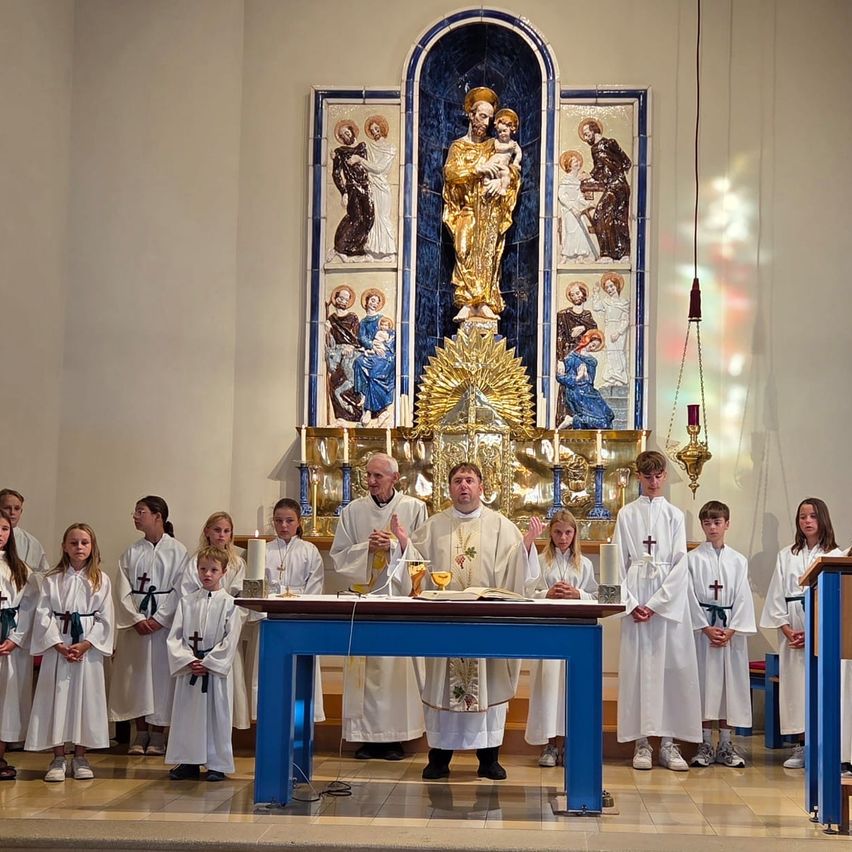 A priest stands at the altar with a chalice and book, surrounded by children in white robes, in a church with a golden statue and mosaics.