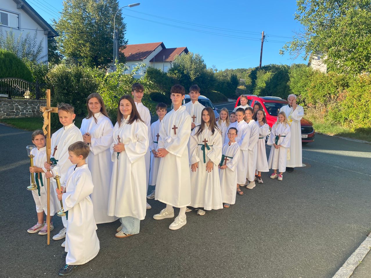 A group of young people dressed in white robes with green sashes and crosses stand in a line outside. They are holding candles and a cross.