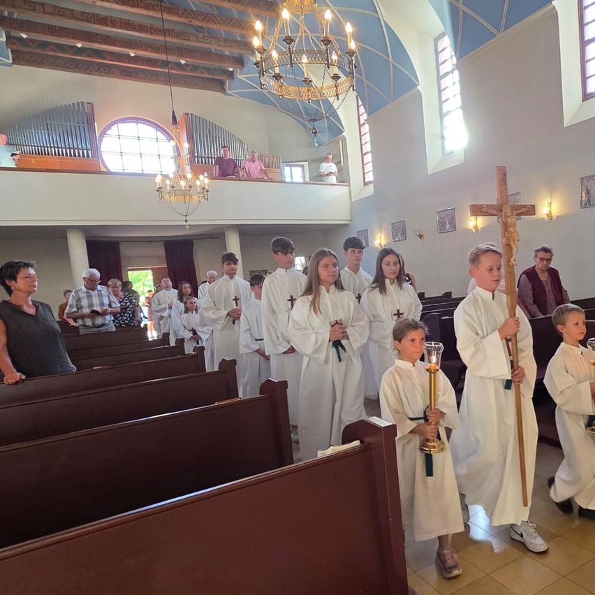 A group of children in white robes holding candles and a cross stand in a church. Behind them, several people are standing and watching.