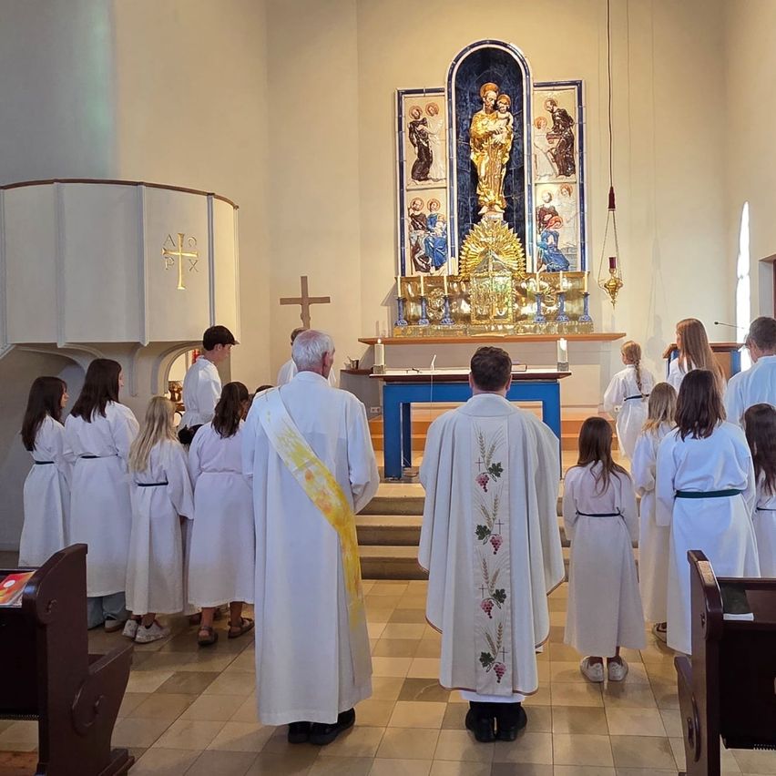 A group of people in white robes are gathered in a church, facing the altar with a golden statue. Priests and altar servers are present.