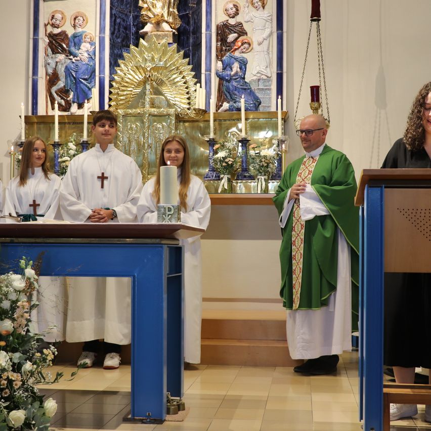 A priest stands before an altar with four young people dressed in white robes, holding candles. Behind them, a golden altarpiece with religious figures is visible.