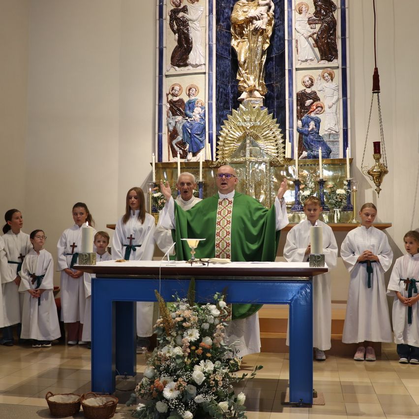 A priest stands at an altar in a church, surrounded by young altar servers. Behind him, a golden statue of a saint is displayed on the altar. Candles and flowers adorn the altar, and the priest is wearing a green and gold vestment.