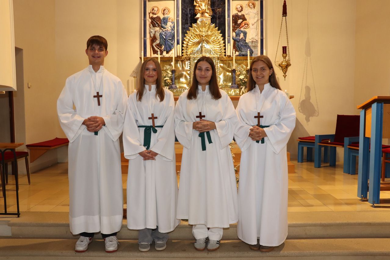 Four individuals dressed in white robes with green sashes and crosses stand in front of a church altar.