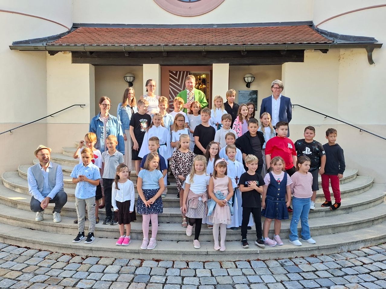 A group of children and adults pose for a photo in front of a building with a tiled roof and lamp posts.