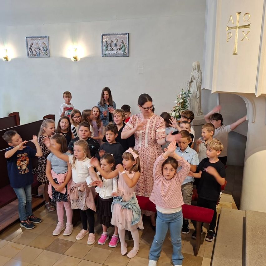 A group of children and adults stand in a church, some with hands raised, in front of a statue of the Virgin Mary.