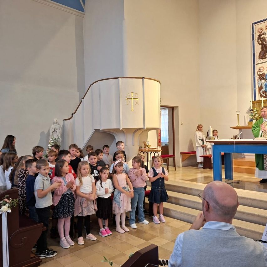 A group of children and adults stand in a church, facing a priest at the altar. The priest is dressed in green and white. The children are wearing various outfits and shoes.