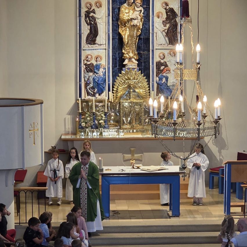 A priest leads a religious service in a church with altar, candles, and a golden statue. Children and adults are present.