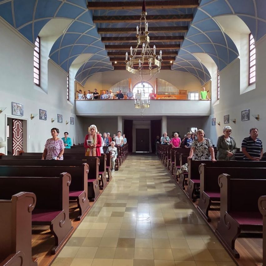 A group of people stand and sit in a church. Wooden pews, a chandelier, and a balcony are visible. The walls have framed pictures.