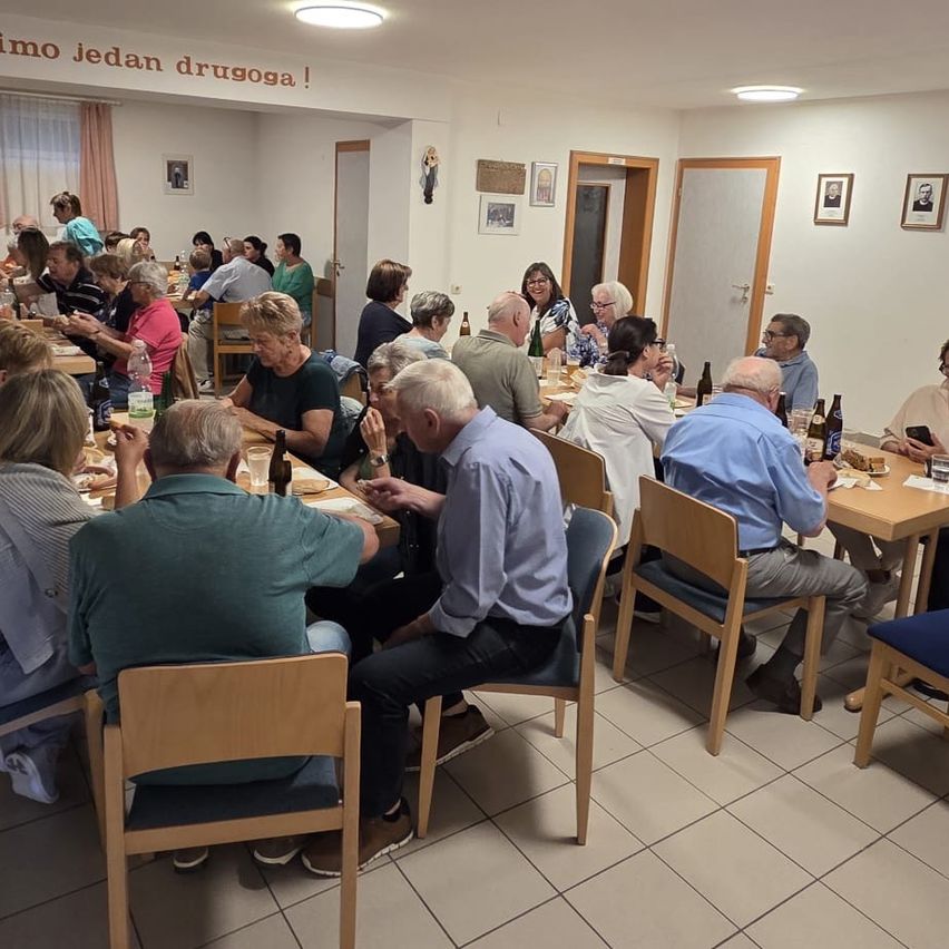 A group of people are sitting around tables in a dining room. Bottles and cups are on the tables. There are picture frames on the walls.