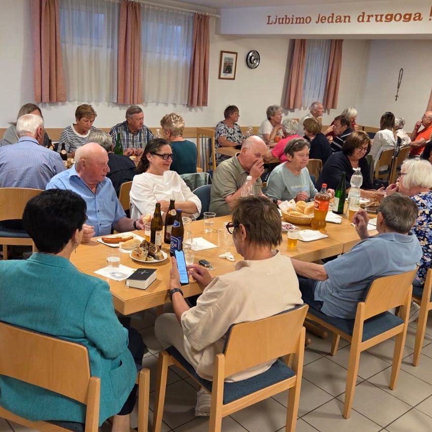 A group of elderly individuals are seated around a table in a dining room, engaging in conversation and enjoying their meal. Various items, including bottles, glasses, and food, are on the table. Above, a wall displays a picture frame and a decorative banner.