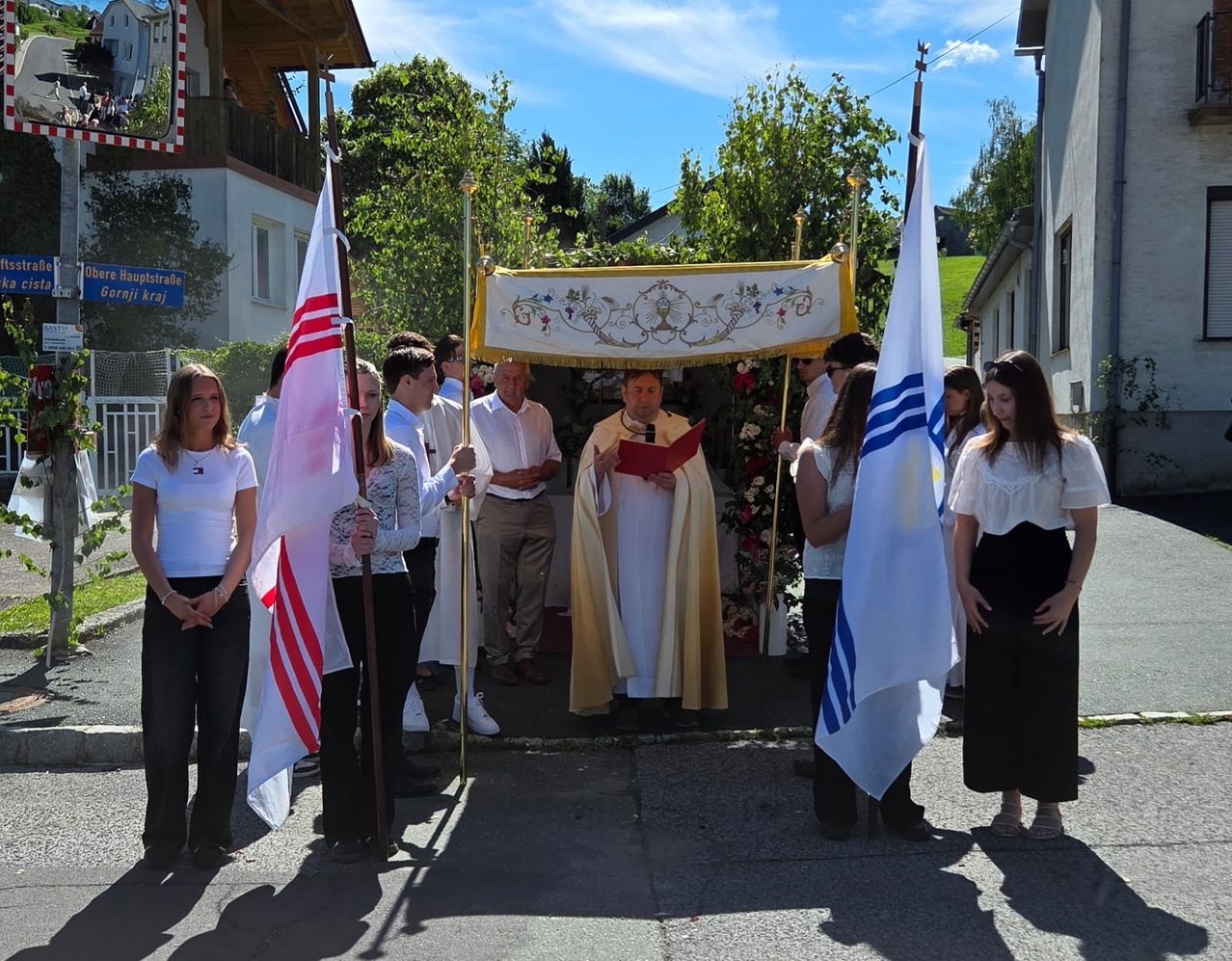 A priest reads from a book while a group of people holding flags stand around him in a courtyard.