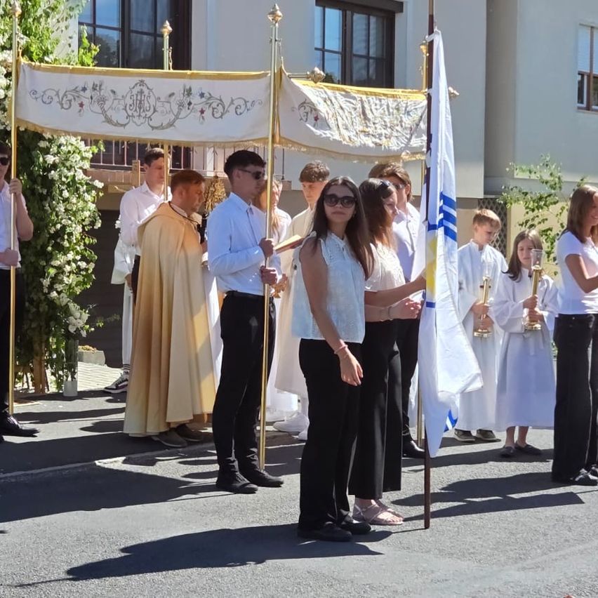 A group of people in traditional attire are gathered in an outdoor setting. Some hold a flag and a book. They stand near a decorative archway.