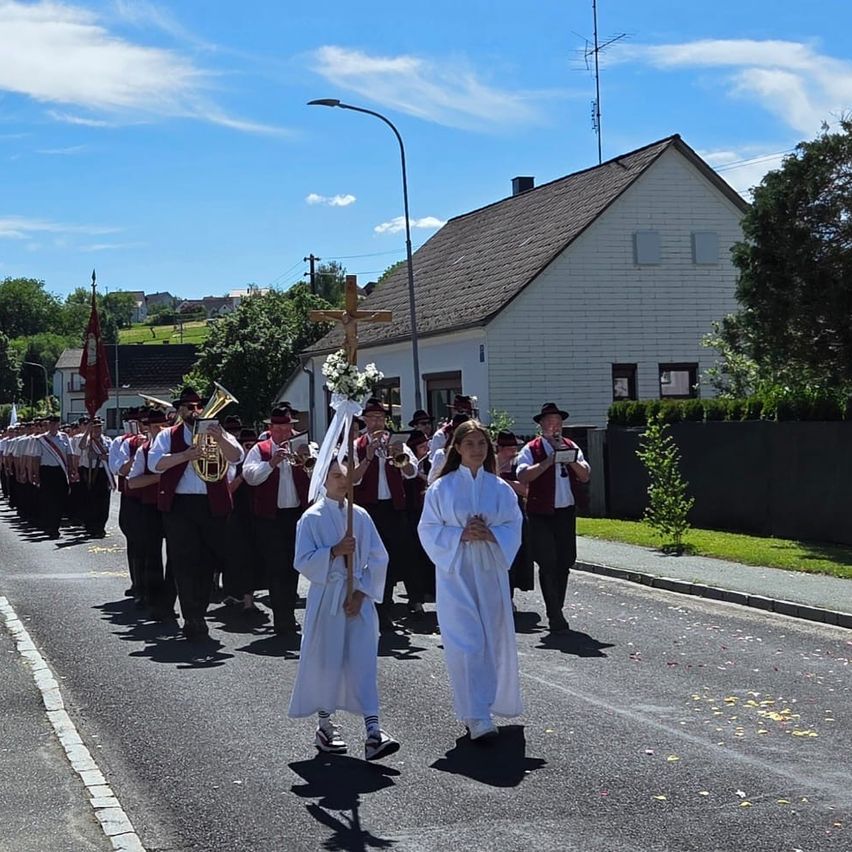 A processional march with children in white robes, a brass band, and a cross in a small town.