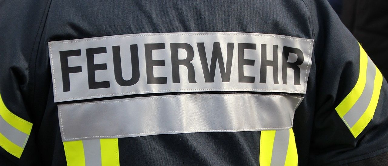 Nahaufnahme der Feuerwehruniform mit dem Wort 'FEUERWEHR' auf dem Rücken. Die Uniform hat reflektierende gelbe und graue Streifen.