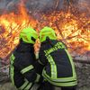 Zwei Feuerwehrleute mit Helmen, auf denen 'TERNITZ-FLATZ' steht, stehen vor einem brennenden Holzhaufen.