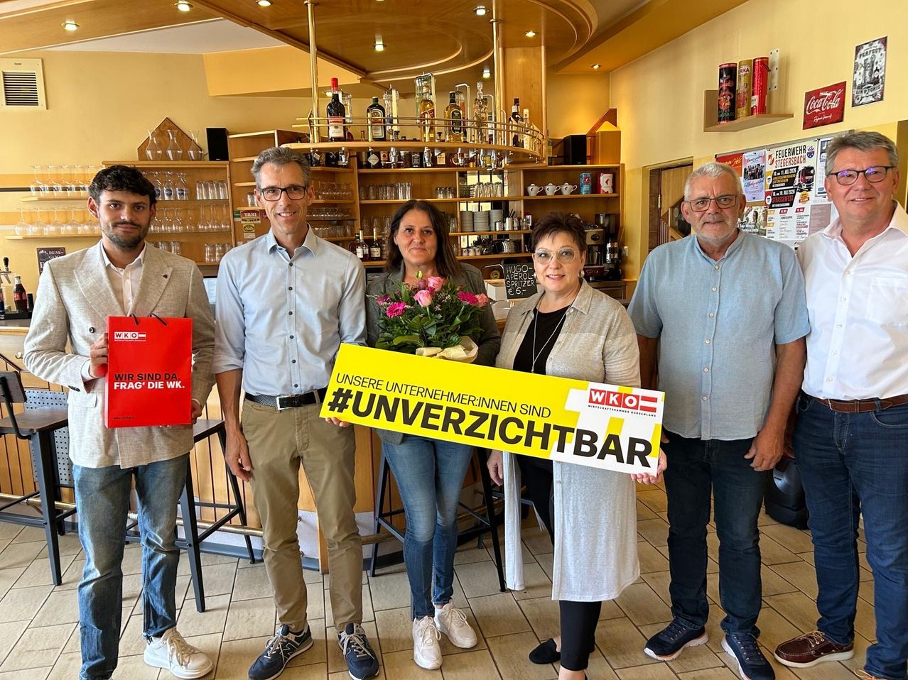 Fünf Personen stehen in einer Bar, lächeln und posieren für ein Foto. Eine Frau hält ein gelbes Schild mit dem Hashtag #unverziichtbar. Hinter ihnen sind Regale mit Flaschen und Gläsern zu sehen.
