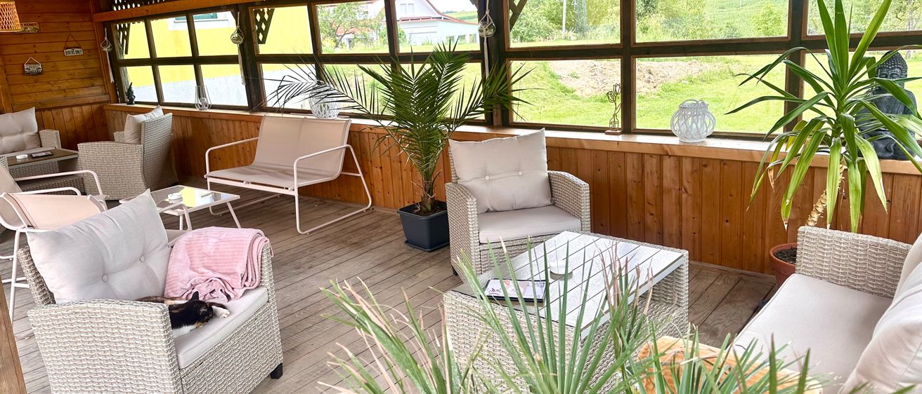 Ein gemütlicher Außenbereich mit weißen Korbmöbeln und Topfpflanzen. Die hölzerne Terrasse ist geräumig, mit großen Fenstern, die einen Blick auf eine grüne Landschaft bieten.