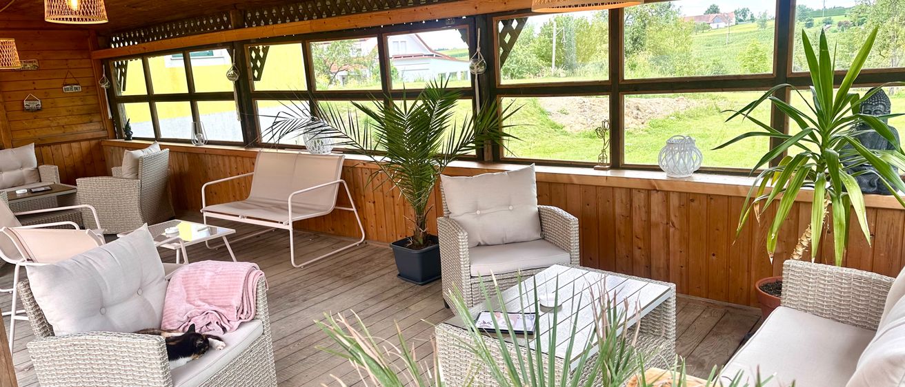 Ein gemütlicher Außenbereich mit weißen Korbmöbeln und Topfpflanzen. Die hölzerne Terrasse ist geräumig, mit großen Fenstern, die einen Blick auf eine grüne Landschaft bieten.
