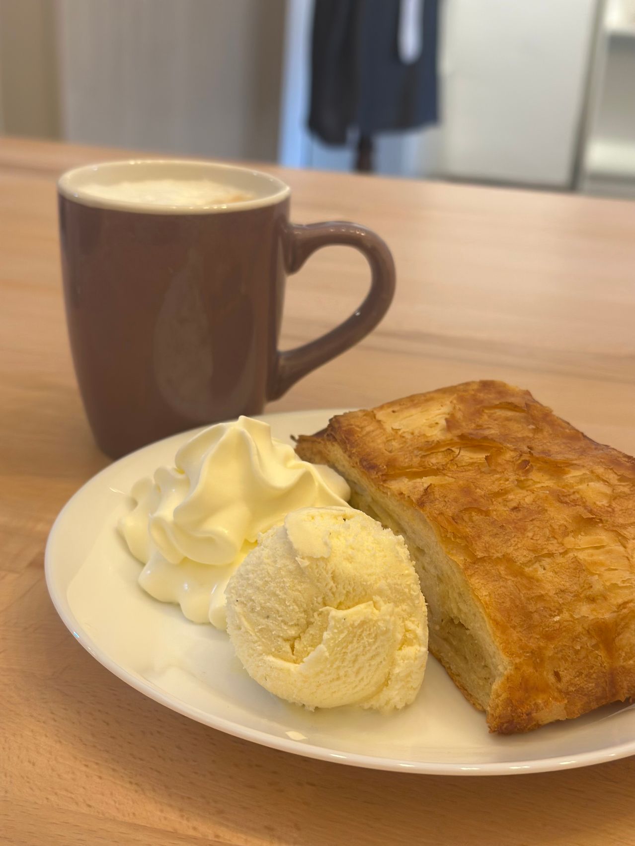 Ein weißer Teller mit Gebäck, Schlagsahne und einer Kugel Eis steht neben einer braunen Kaffeetasse auf einem Holztisch.