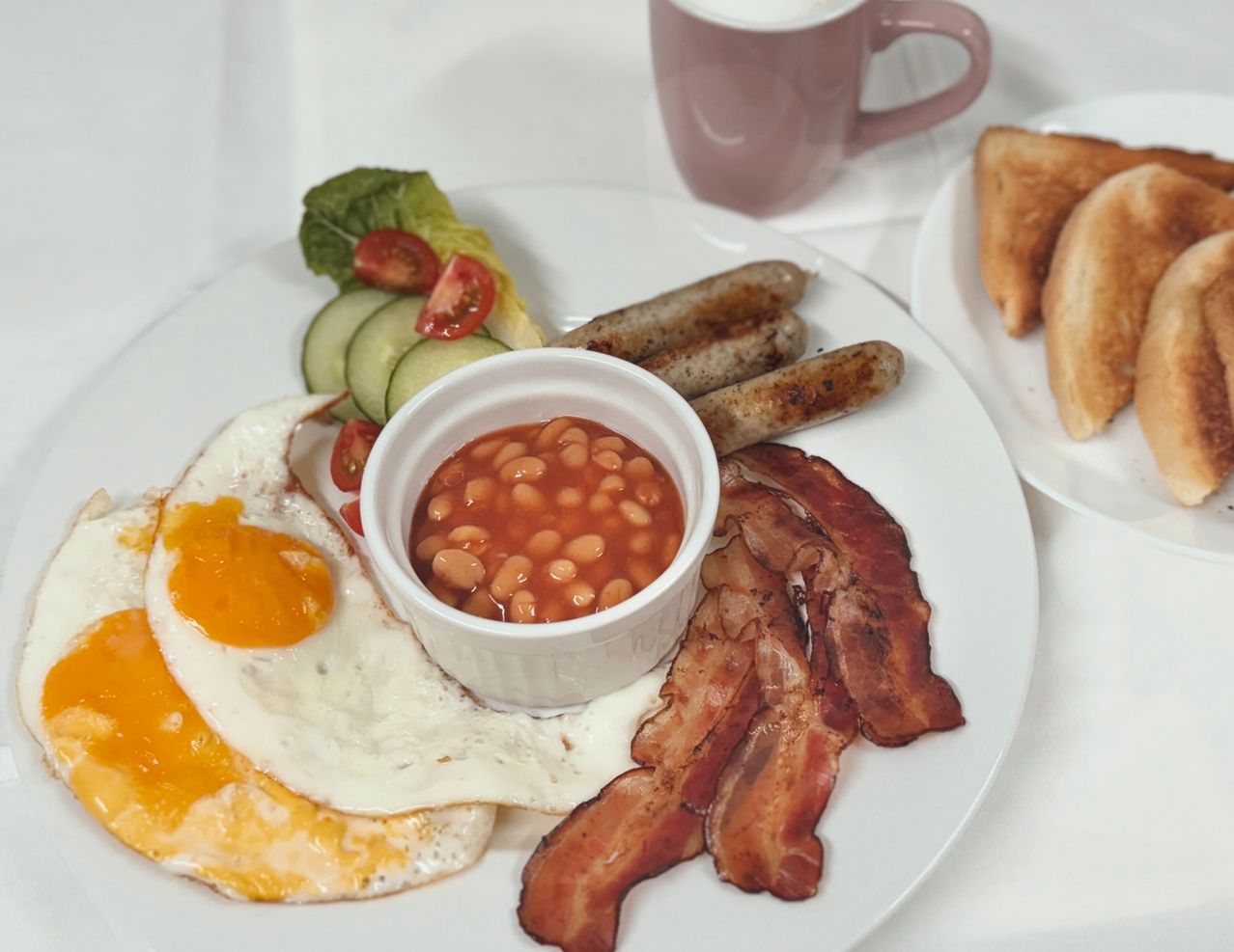 Ein Frühstücksteller mit gebratenen Eiern, knusprigem Speck, Würstchen, gebackenen Bohnen, Gurkenscheiben und Kirschtomaten. Ein rosafarbener Becher und Toastbrot sind ebenfalls vorhanden.