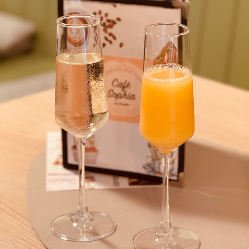 Zwei Weingläser stehen auf einem Tisch, eines gefüllt mit einer rosa Flüssigkeit und das andere mit Orangensaft, vor dem Hintergrund einer Cafe Sophia Speisekarte.
