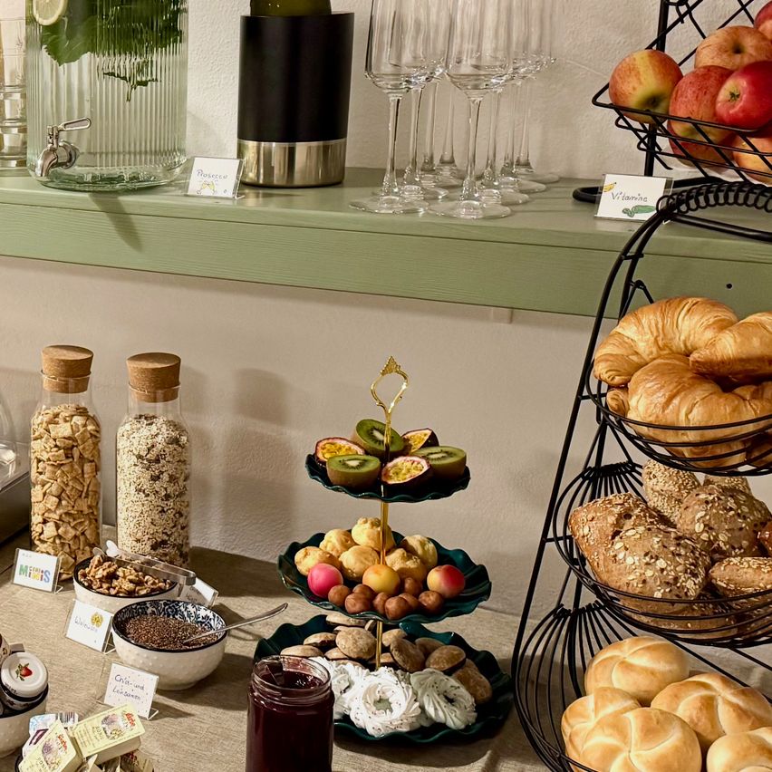 Ein Frühstückstisch mit Croissants, Brot, verschiedenem Obst und Desserts auf einem dreistufigen Ständer. Ein Glas Marmelade, Müsli und Nüsse sind ebenfalls auf dem Tisch.