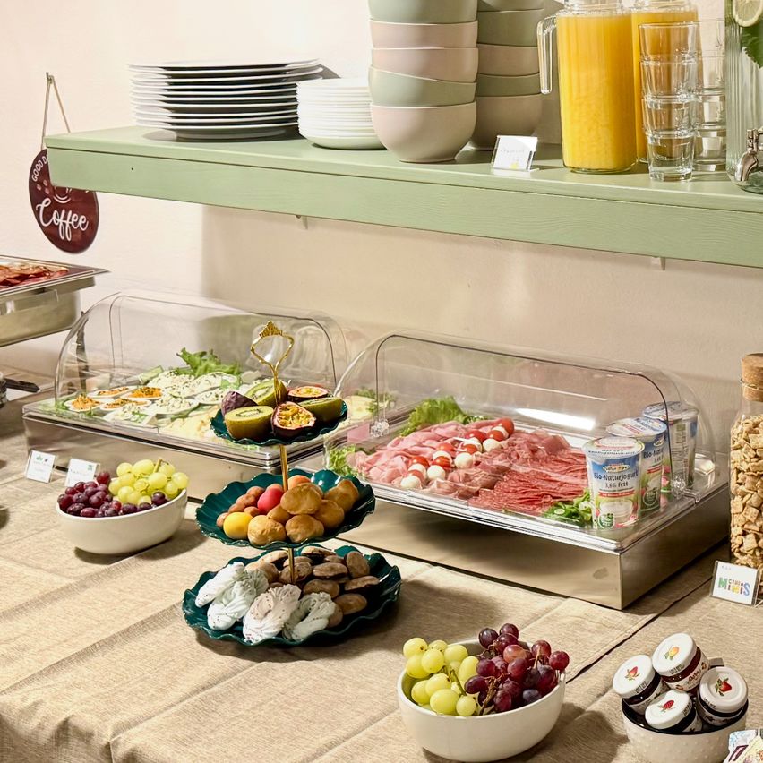 Ein Buffettisch zeigt verschiedene Speisen, darunter Salate, Fleisch, Obst und Desserts. Teller, Schüsseln und Gläser stehen auf einem Regal darüber. Getränke werden in Gläsern serviert.