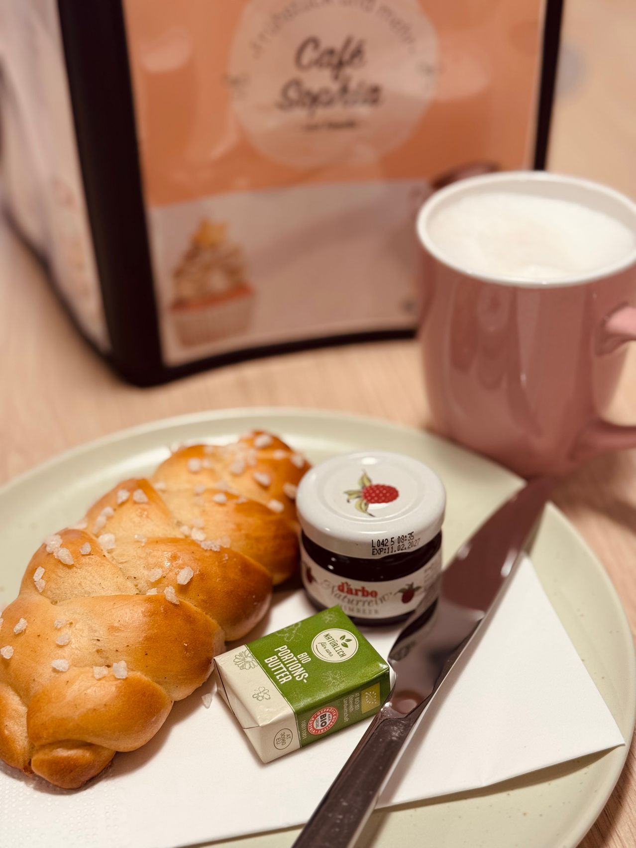 Ein Teller mit Brezeln, einem Messer, einem Glas Marmelade und einer rosa Tasse mit einem Getränk. Im Hintergrund ist ein verschwommener Bäckereibeutel zu sehen.