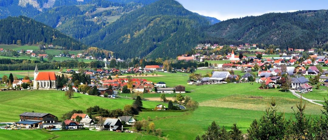 Ein malerisches Dorf in einer bergigen Region, umgeben von üppigen grünen Feldern und dichten Wäldern. Die Häuser, einige mit roten Dächern, liegen am Fuße eines Hügels unter blauem Himmel.