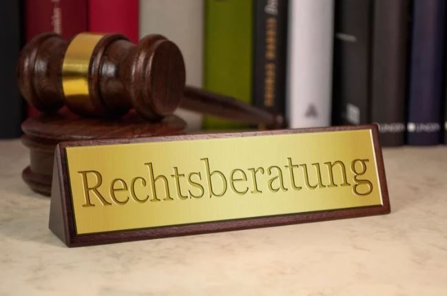 Ein hölzerner Richterhammer liegt auf einem Schild, das 'Rechtsberatung' in goldenen Buchstaben vor einer Reihe von Büchern zeigt.