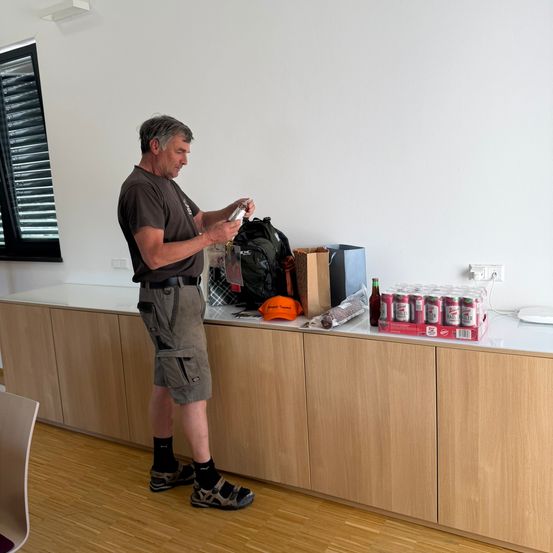 Ein Mann steht in einem Raum, hält eine Flasche und hat einen Rucksack und Gegenstände auf einer Theke hinter sich.