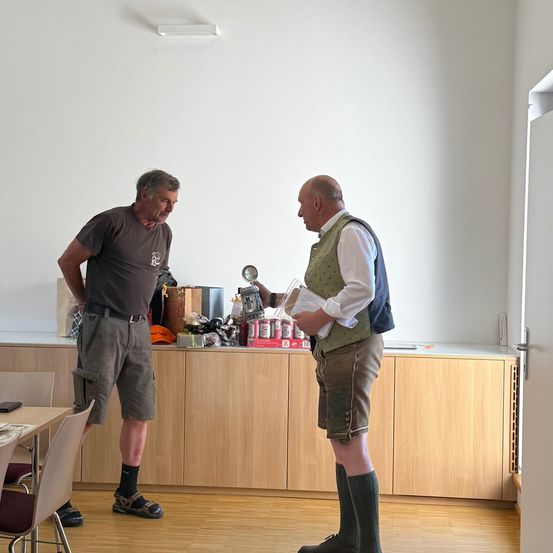 Zwei Männer stehen in einem Raum mit Holzböden und Schränken, einer hält eine Trophäe und Papiere, der andere sieht ihn an, mit Gegenständen und einer Tasche hinter ihnen.